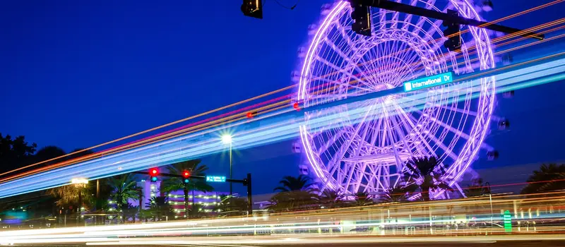 The Orlando Eye observation wheel lit up purple at night on International Drive, with light trails from passing traffic