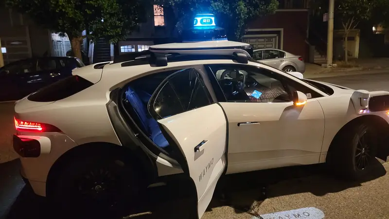 A Waymo autonomous Jaguar I-Pace at night, white car with roof sensors lit under street lights